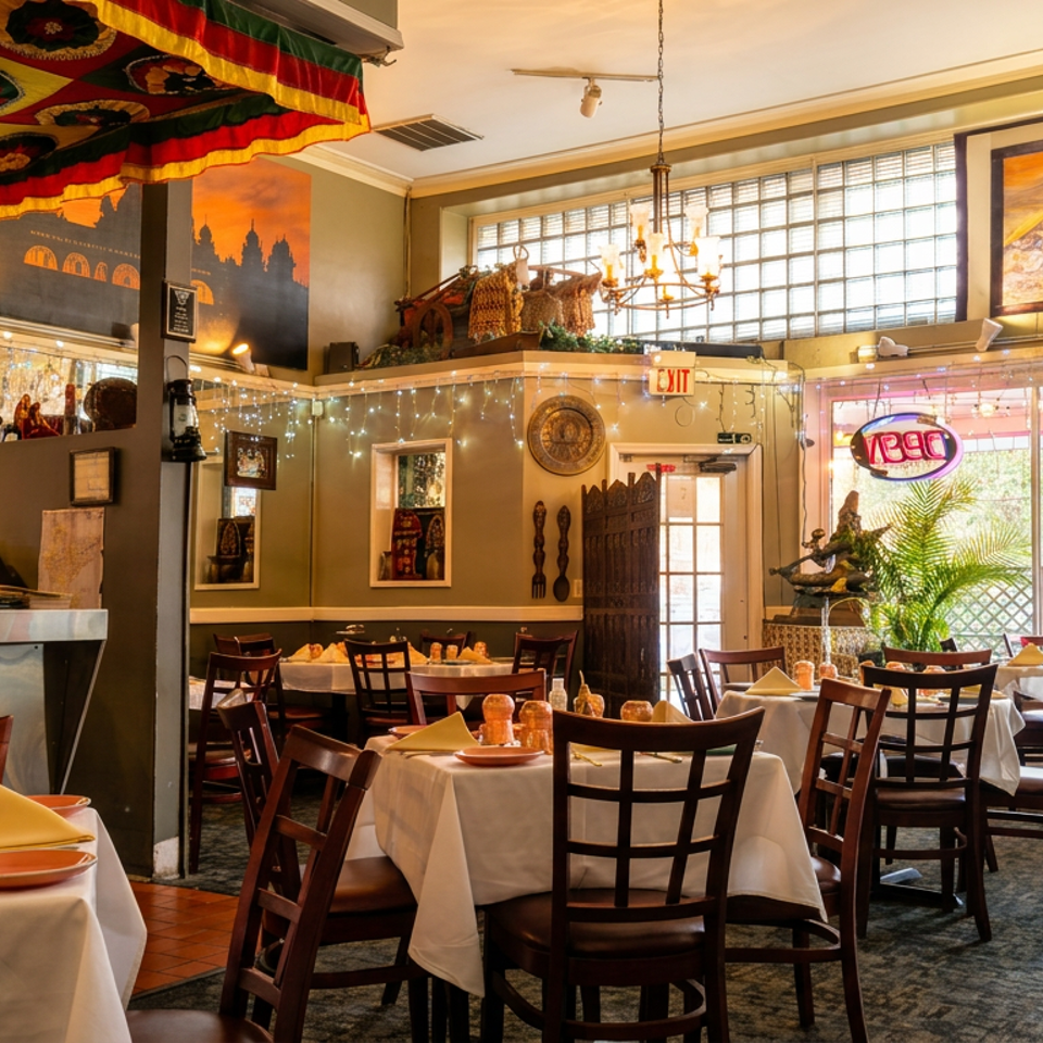 Colorful restaurant dining room with white-tablecloth tables, dark wood chairs, bar, chandeliers, artwork, and bright windows.